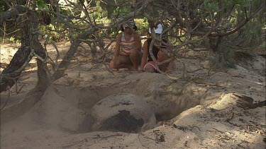 Two girls sit next to a green turtle. Green turtle digs a pit for laying its eggs.