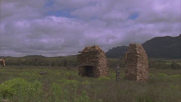 ranges in background and dilapidated ruins of old homestead in foreground and horse riders ride towards stone remains