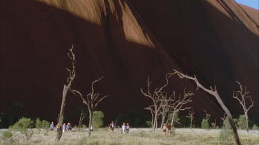 pan across Uluru rock in background with shadow cast and people walk at base