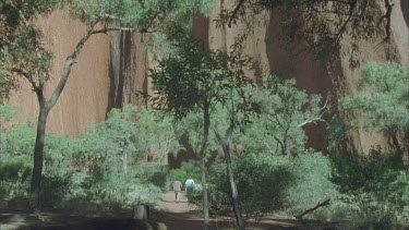 tilt up from tourists on ground to perimeter of gorge with trees in foreground and rock of Kata Juta some blue sky behind