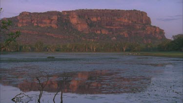 Nourlangie Rock and escarpment from Nawurlandja Lookout vista with water in foreground and reflection of rock in water *