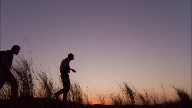 silhouette shot of Klaus Kruiper tracking walking across crest of hill with grass tussocks followed by Louis Liebenberg