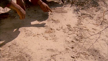 Bushman pointing to tracks in earth with fingers then stand up and walk out of shot
