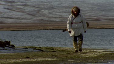 frozen waters edge of a lake, pan up to MWS of tundra landscape, Inuit man walking towards camera.