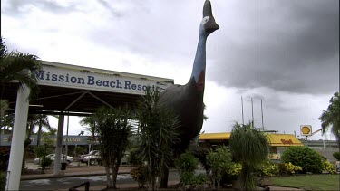 Establishing shot of Mission Beach Resort with giant Cassowary statue out front. Garage on rhs. Dark clouds in sky.