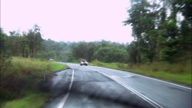 In a car driving through rain. The windscreen wipers wipe across the windscreen. Tropical landscape in background, palm trees and lush green vegetation, a rural scene.