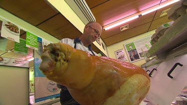 Cut of meat in butcher's shop. Butcher in traditional blue apron slices meat of leg of animal (pig?)