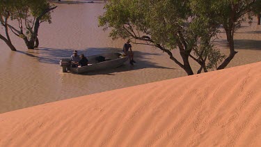 Three people fishing in tinny boat with outboard motor. They are on very shallow waters of flooded desert plain. High Angle. In foreground Big Red famous sand dune.