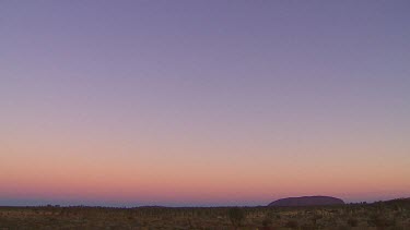 Big wide shot, establishing shot. Uluru at sunset. The horizon has layers of colours, purple, pinks and range hues. Uluru in distance in silhouette.