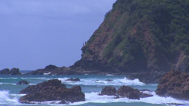 Two people on precarious cliff ledge looking at surf waves crashing to cliffs below.