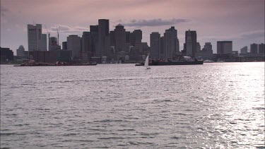 Sailingboat sailing slowly on in front of Boston skyline on a cloudy day, from right to left. Two larger boats behind the sailingboat.