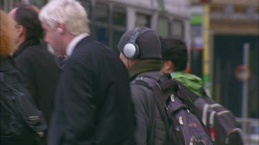 Pedestrians walking across street. Business people and young people listening to music. Busy street scene in city. Bus drives past.