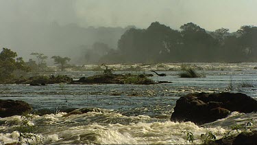 Victoria Falls waterfall. Pan of Zambezi River standing on the lip of the falls. Famous Victoria Falls bridge can be seen through the mist.