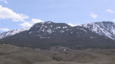 Forested snowy mountains, blue sky, clouds and grassy hills