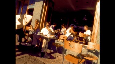 Sepia. Exterior. People drinking coffee at a Bondi cafe. People with surf boards walk past.