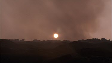 Apocalyptic landscape. Steam and smoke blowing across screen. Dark volcanic rocky landscape. Eerie. The sun is setting, a red ball just above the horizon.