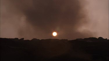 Apocalyptic landscape. Steam and smoke blowing across screen. Dark volcanic rocky landscape. Eerie. The sun is setting, a red ball just above the horizon.