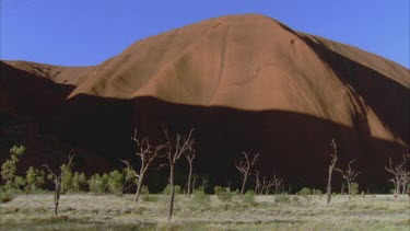 pan across Uluru rock in background with shadow cast and people walk at base