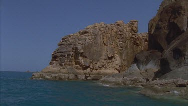 tracking along island shore with large boulders at shore and yacht appears in anchorage