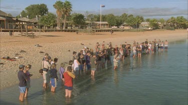a crowd of tourists stand on beach watching dolphin in shallow water