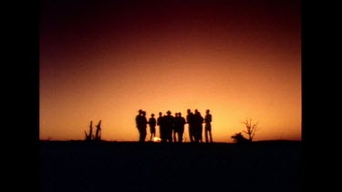 Zoom in. Men dancing around campfire in silhouette against red and orange sunset sky.