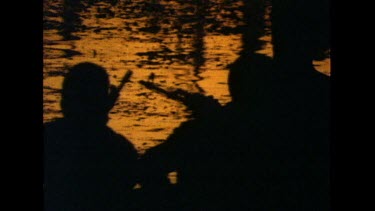 Men in canoe. Silhouette against red orange sunset reflected on water.
