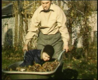 Father wheels a toddler in a wheelbarrow filled with autumn maple leaves