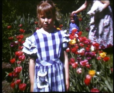 Girl stands in patterned tartan dress beside colourful flowers, father mows garden and a cat attacks a toodler