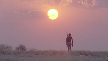 bushman walking towards camera with sunburst sunset behind , with bow and arrows over shoulder , very nice shot , a classic , very steady , various takes
