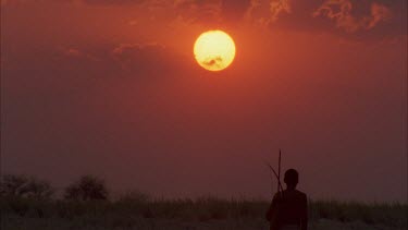bushman walking towards camera with sunburst sunset behind , with bow and arrows over shoulder , very nice shot , a classic , very steady , various takes