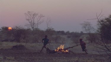 Bushman camp , with campfire a slight pink glow in the sky , dusk with young boys playing around campfire