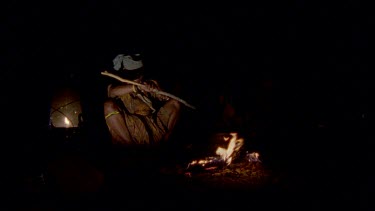 Bushman camp , with campfire , woman sitting beside fire , smoking a pipe , stirring cooking pot takes pot off fire