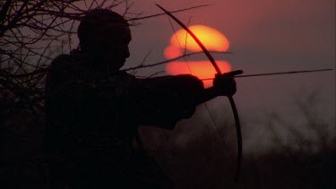bushman fires arrow from bow against sunset