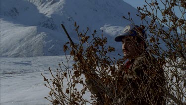 Inuit hunter with a gun. Shoots from behind a tree, walks out of screen off into the snow