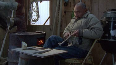 Inuit man carving from walrus ivory in shed or hut with tools in background