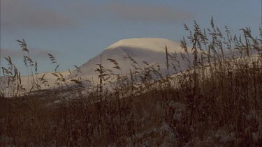 pan across Grass peaking through snow covered ground, still water, snowy mountain in background