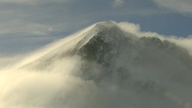 Wind blows strongly covering mountain peak summit in cloud. Shot shows cloud, mist travelling up the mountain. The clouds are brightly illuminated by the sunshine behind. Shows how wild weather can oc...