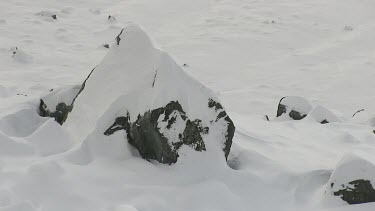 Thick snow and ice on mountain peak, slow pull back reveals lake with thin layer of thawing ice