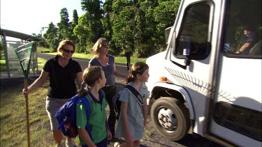 Mothers armed with rakes walk their children along the road. They are carrying the rakes to fend off potential attack by cassowary. Children get into school bus.
