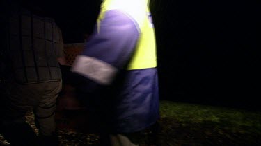 Two Park rangers at night. It is raining. They are lit by the headlamps of a car. They carry a crate, inside it is a cassowary that is being relocated into rainforest area.