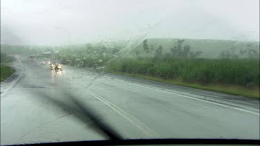 In a car driving through rain storm. The windscreen wipers wipe across the windscreen. Tropical landscape in background, palm trees and lush green vegetation, a rural scene.