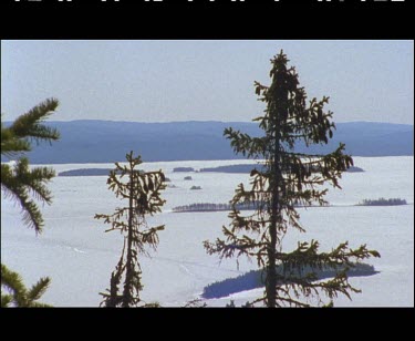 Wide Shot of winter landscape. Wide snow fields and tips of conifer trees. Frozen lake . Matching shot of this lake in spring AWVsc-1061