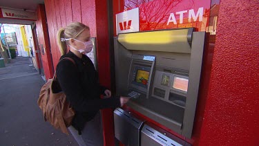 Woman wearing medical face mask approaches suburban cash teller machine to withdraw money