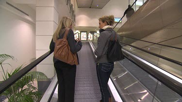 Two women ride up escalator in shopping mall