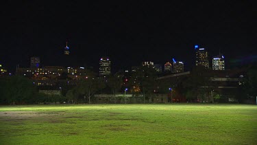Skyline of the city at night from floodlit green sports field, empty.