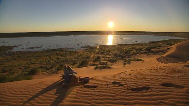 Sun setting over desert flood plain, person in foreground in silhouette lying on red sand dune relaxing