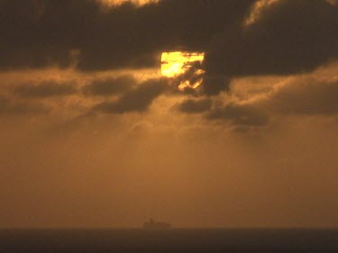 Red orange sunset. Sun is a yellow red ball obscured behind clouds. Ship on horizon. Calm ocean.