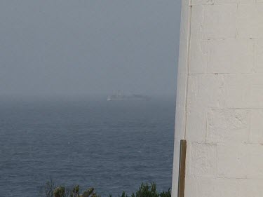 Container ship on horizon, lighthouse in foreground, heavy white wall framing the ocean in background.