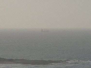 Fishing boat or ship at sea waves and rocks in foreground. Sky is gloomy or dusty makes image unclear