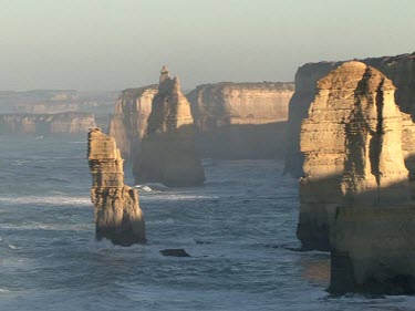 Twelve Apostles, rock stacks. Limestone eroded by wave action. Sun shining and glistening on water. See back of one woman's head as she looks out to sea. Her hair is blowing in the wind.